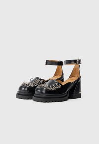 Pantofi cu toc chunky din piele vernis neagră, cu brățară la gleznă și detalii din lanț metalic pe partea superioară. Talpă din cauciuc texturat.
