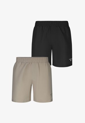 Homme musclé avec de nombreux tatouages sur le torse, les bras et les jambes, portant un short beige et des colliers argentés, debout pieds nus sur un fond gris.