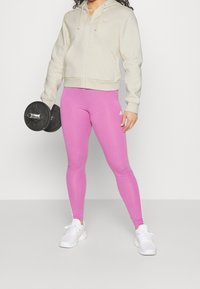 Osoba oblečená v krémovém zipovém mikině a růžových legínách Adidas, držící černý činkový závaží v pravé ruce, stojící na pozadí s jednobarevným vzorem.