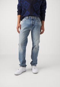 Helle blaue Denim-Jeans mit einem lässigen Schnitt, die eine subtile Ausbleichung aufweisen. Getragen mit weißen Sneakers, präsentiert vor einem schlichten Hintergrund.