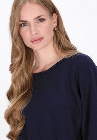Granatowy sweter z szerokim dekoltem, miękką teksturą i długimi rękawami. Modelka ma długie, falujące włosy oraz złote kolczyki w kształcie kółek.