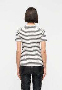 Γυναίκα με ίσια καρέ κουπ, που φοράει λευκό μπλουζάκι με κοντά μανίκια και μαύρες οριζόντιες ρίγες και μαύρο δερμάτινο παντελόνι, που στέκεται γυρισμένη προς τα πίσω.