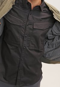 Camicia nera con bottoni, dotata di due tasche sul petto, realizzata in tessuto liscio, indossata sotto una giacca verde aperta con fodera grigia.