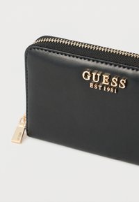 Fekete bőr pénztárca aranyszínű kiegészítőkkel, cipzáras záródással és az előlapján dombornyomott "GUESS EST 1981" logóval.