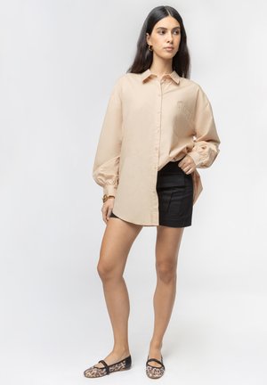 Donna con lunghi capelli scuri che indossa una camicia beige oversize con bottoni, una minigonna nera, ballerine con stampa leopardata e orecchini a cerchio dorati.
