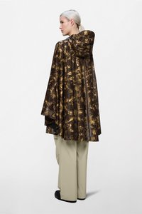 Καφέ και χρυσή ποδιά με σχέδιο και κουκούλα, κατασκευασμένη από γυαλιστερό, αδιάβροχο υλικό. Φορεμένη πάνω από μπεζ παντελόνια και μαύρα παπούτσια.