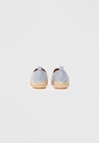 Gaiši zili teksturēti espadrilles ar pītām jutu zoli, redzami no aizmugures uz vienkārša balta fona.