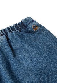 Modrá džínová tkanina s elastickým pasem a malým hnědým vyšitým logem poblíž horního okraje na bílém pozadí.