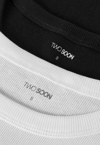 Twee T-shirts, één zwart en één wit, met een geribbelde textuur getoond. Labels met de tekst "TWO SOON" en maat "S" in contrasterende kleuren.
