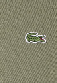 Tissu vert olive avec un tissage texturé, présentant un petit logo de crocodile vert brodé avec des accents rouges et un contour blanc.