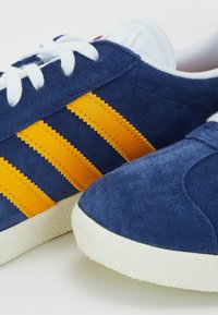 Blå mockasneakers med tre gula ränder, vita skosnören och en texturerad gummisula. Har en rundad tå och kontrasterande detaljer.