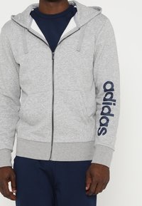 Veste à capuche zippée grise en matériau doux, avec un grand logo "adidas" en bleu marine sur la manche gauche et une capuche doublée.