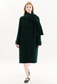 Vihreä villakangas takki, jossa on suuri käännekaulus, kaksi etutaskua ja pitkät hihat nappikoristeilla. Mustat kengät alla.