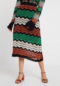 Kvinna som bär en midi kjol med zickzackmönster i grönt, brunt, svart och vitt samt en matchande långärmad topp med bruna klacksandaler.