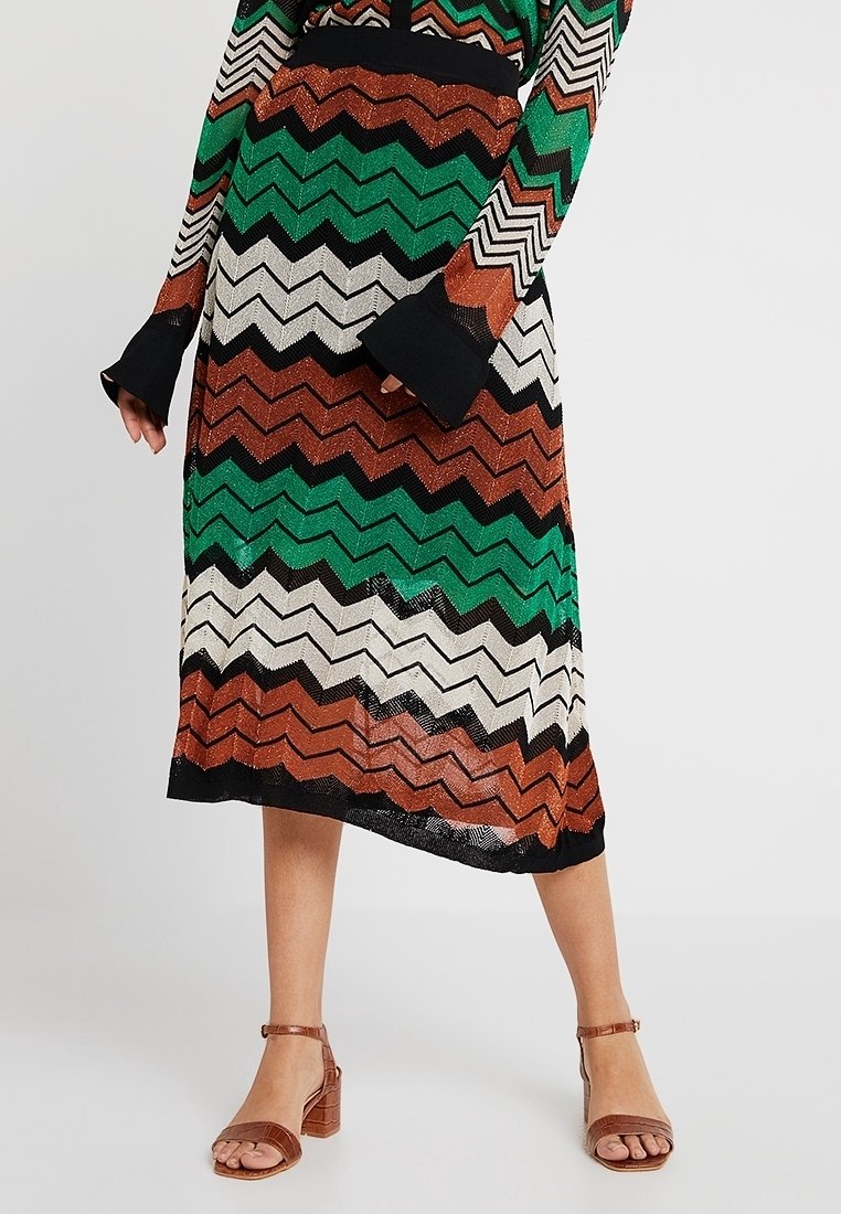 Kvinna som bär en midi kjol med zickzackmönster i grönt, brunt, svart och vitt samt en matchande långärmad topp med bruna klacksandaler.