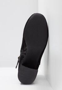 Chaussure noire avec une tige en mesh texturé, un bout arrondi et une semelle plate en caoutchouc avec un motif côtelé. Détail zippé sur le côté.