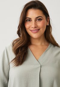 Lichtgroene blouse met een v-hals, zachte textuur en subtiele plooidetails. Draagt met lang, golvend haar. Eenvoudig ontwerp, zonder patronen.