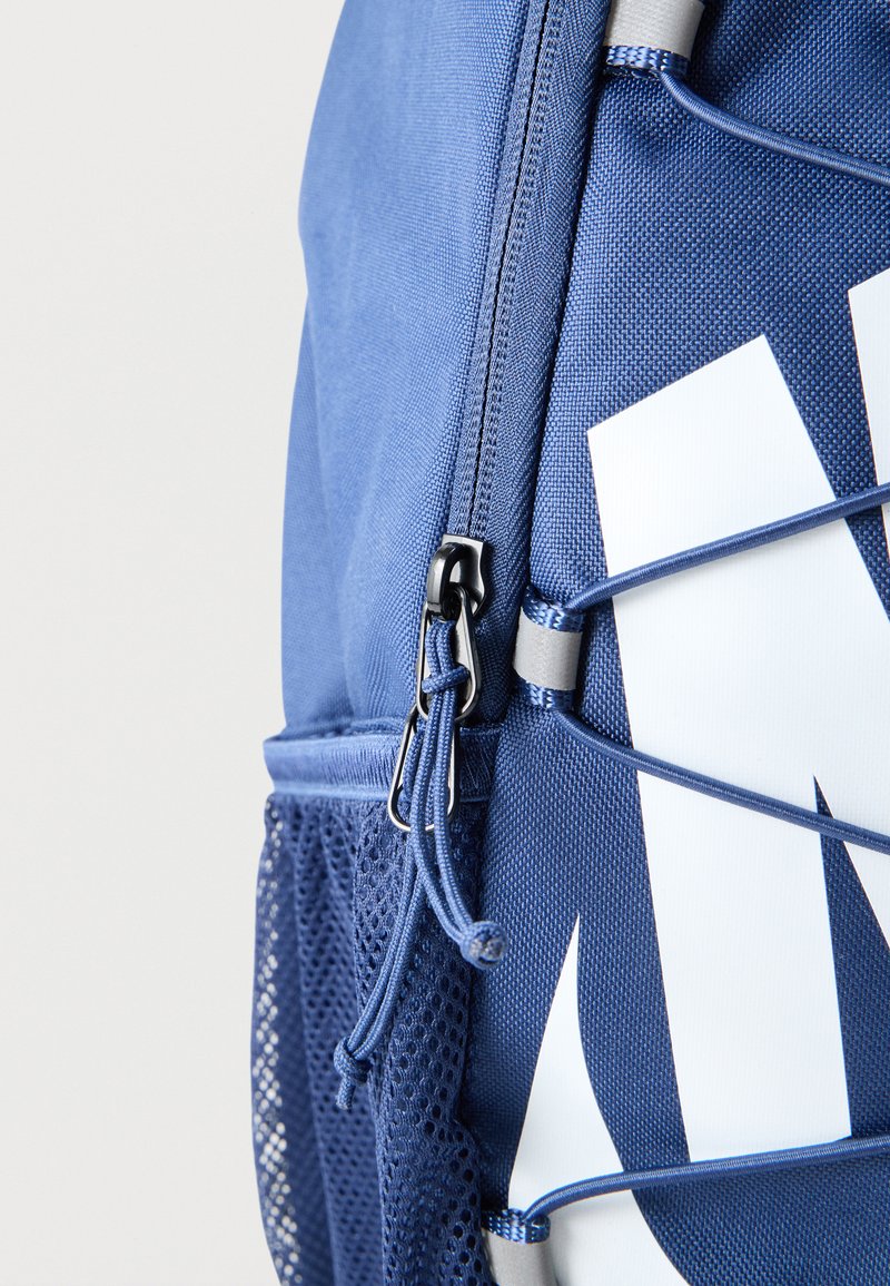 Nahaufnahme des seitlichen Reißverschlusses, des Mesh-Fachs und des weißen Swoosh-Logos mit elastischer Kordeldetailierung an einem blauen Nike-Rucksack.
