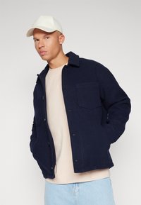 Marineblå uldblandingsjakke med krave, for- lommer og knaplukning, båret over en lys beige skjorte. Model iført en hvid kasket.