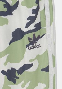 Acercamiento de una tela de camuflaje verde, blanca y negra con el logo negro de Adidas y detalles en blanco en la prenda.
