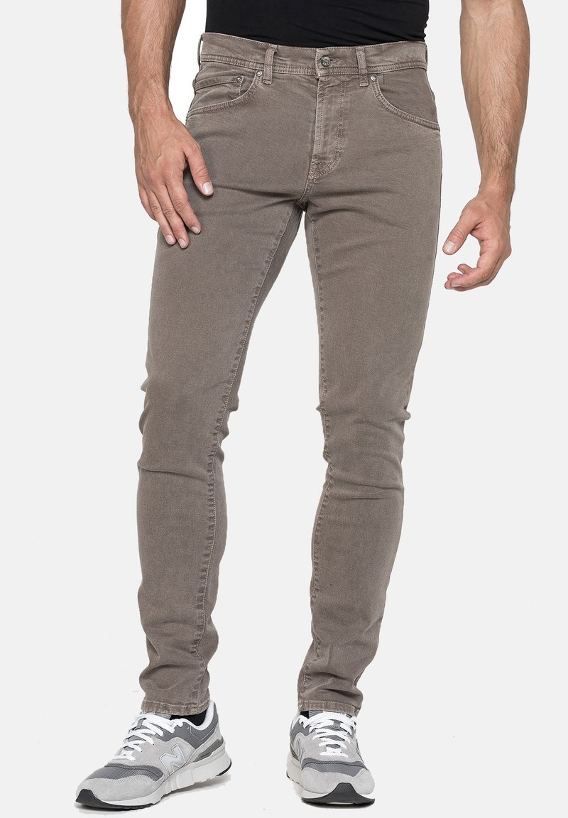 Jeans in cotone marrone slim-fit con una trama sottile. Le caratteristiche includono cinque tasche, chiusura a bottone e cuciture a contrasto. Indossati con sneakers grigie.
