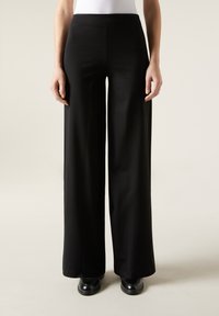 Pantalons noirs à larges jambes en tissu lisse, avec un design minimaliste et une taille haute, associés à des chaussures noires.