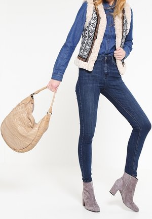 Mujer con conjunto de mezclilla azul, chaleco con ribete de piel estampada, botines de ante gris, sosteniendo un bolso grande beige por el asa.