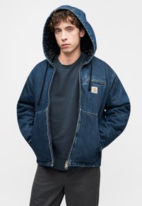 Τζιν μπουφάν με κουκούλα και φερμουάρ, σκούρο μπλε χρώμα, ριμπ μανσέτες και τσέπη στο στήθος με ετικέτα λογότυπο. Φορεμένο πάνω από ένα σκούρο φούτερ.