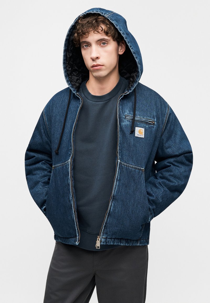 Denim hættejakke med lynlåslukning, mørkeblå farve, ribbet manchetter og en brystlomme med en logomærkat. Båret over en mørk sweatshirt.