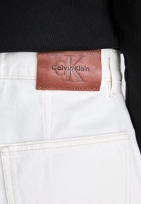 Witte denim jeans hebben een tan leren label met "Calvin Klein" in zwart gegraveerd, gelegen bij de tailleband. Een gestructureerde zwarte shirt is zichtbaar erboven.