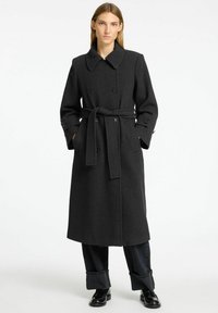 Dolga črna plašč v herringbone vzorcu z dvojnim žepkom na sprednji strani, velikim ovratnikom, pasom v pasu in stranskimi žepi.