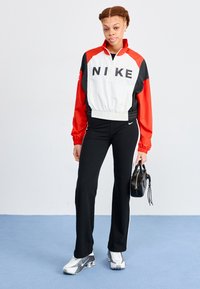Nike vēja jakas ar baltu, sarkanu un melnu dizainu, ar ceturtdaļas rāvējslēdzēju, elastīgu jostasvietu un izteiksmīgu logo. Apvienots ar melnām biksēm un sporta apaviem.