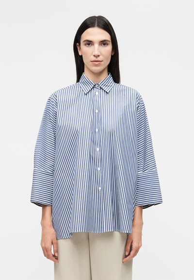 Femme aux cheveux raides et foncés portant une chemise ample à rayures verticales bleues et blanches et un pantalon beige clair.