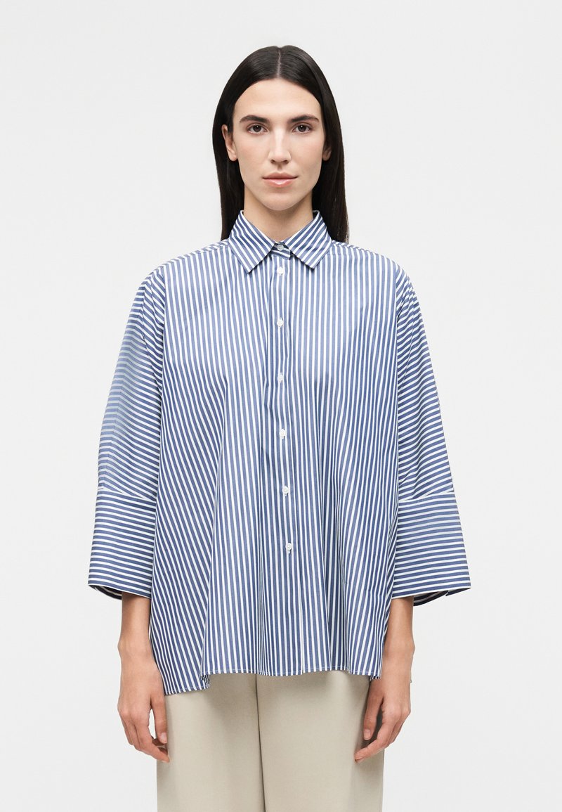 Γυναίκα με ίσια, σκουρόχρωμα μαλλιά που φοράει μια φαρδιά μπλε και λευκή πουκάμισο με κάθετες ρίγες και ανοιχτό μπεζ παντελόνι.