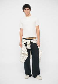 Young man with curly hair wearing a white t-shirt, black wide-leg pants, beige belt, white sneakers, and a patterned jacket tied around the waist.