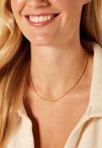 Collier en chaîne torsadée dorée, finement texturé avec un éclat subtil, reposant sur une peau nue, complétant un haut léger et texturé.