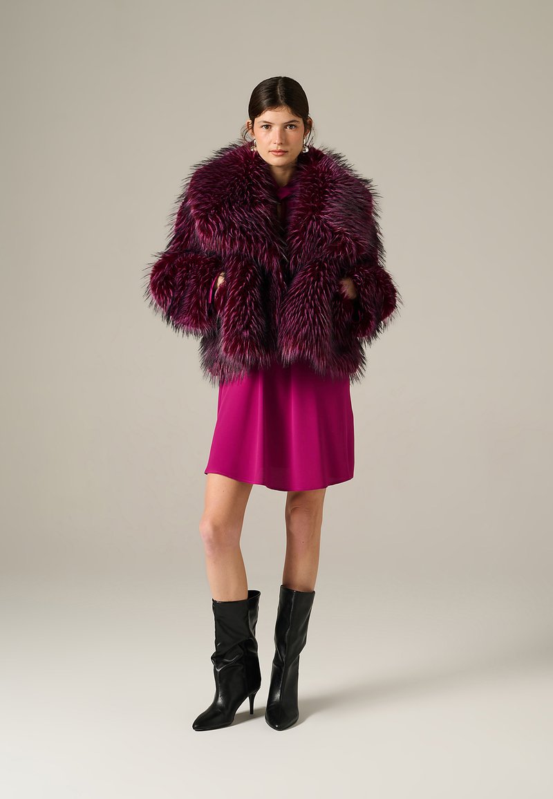 Giacca in pelliccia sintetica di un profondo bordeaux con un design voluminoso, abbinata a un vestito fucsia e stivali neri fino al ginocchio. Texture liscia e lucida.