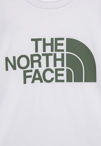 Bílý bavlněný tričko s velkým zeleným logem, které zní "THE NORTH FACE", s tučným písmem bez patkového písma a designovým prvkem ve tvaru čtvrtkruhu.