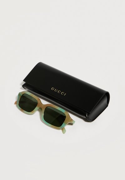 Lunettes de soleil carrées translucides vertes avec des verres foncés à côté d'un étui Gucci noir avec inscription dorée sur une surface blanche.