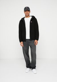 Mustat fleece-tyyppiset vetoketjulliset takit harmaalla vuorilla, valkoinen t-paita, harmaat farkut ja valkoiset lenkkarit. Takissa on pieni logo.