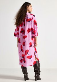 Lang lyserød kjole med rødt blomsterprint, plisseret tekstur og sideslids. Båret med mørkebrune knælange støvler.