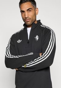 Μαύρο φούτερ με φερμουάρ και λευκές ρίγες στους ώμους, με κεντημένο λογότυπο και υφή με ανάγλυφη όψη.