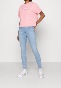 Camiseta corta rosa con un logo en la parte delantera, combinada con jeans ajustados azul claro y zapatillas altas blancas. Textura suave de algodón.