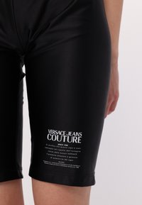 Černé cyklistické šortky s hladkým, lesklým povrchem. Na levé noze mají bílé logo "Versace Jeans Couture" a informace o produktu.