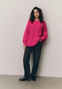 Pulover tricotat supradimensionat de culoare roz intens, cu textură ribbed, purtat cu blugi de denim închis și pantofi slip-on negri și lucioși. Prezentare de ținută casual.