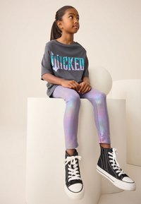 Grå t-shirt med "WICKED" i iriserende palietter, kombineret med skinnende lavendelleggings og sorte høje sneakers med hvide såler.