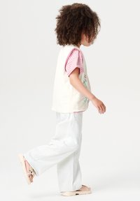 Een kind in een lichtgewicht crème vest met borduursel over een roze en witte gestreepte shirt, gecombineerd met lichte spijkerbroeken en lichte sandalen.