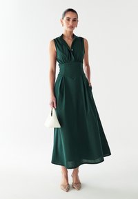 Grünes ärmelloses Kleid mit einem geknöpften Kragen, figurbetontem Bund, plissiertem Rock und seitlichen Taschen, kombiniert mit einer weißen Handtasche und gemusterten Absätzen.