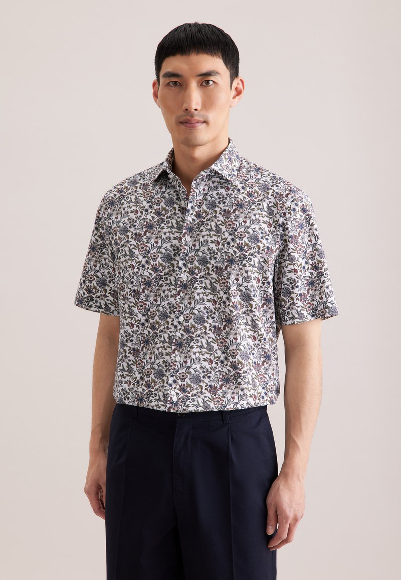 Homme portant une chemise à manches courtes avec un motif floral multicolore et un pantalon bleu marine foncé, sur un fond uni.