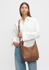 Sac en cuir marron porté sur l'épaule, avec une texture lisse, une forme décontractée et une bandoulière ajustable, tenu par une personne en chemise blanche.
